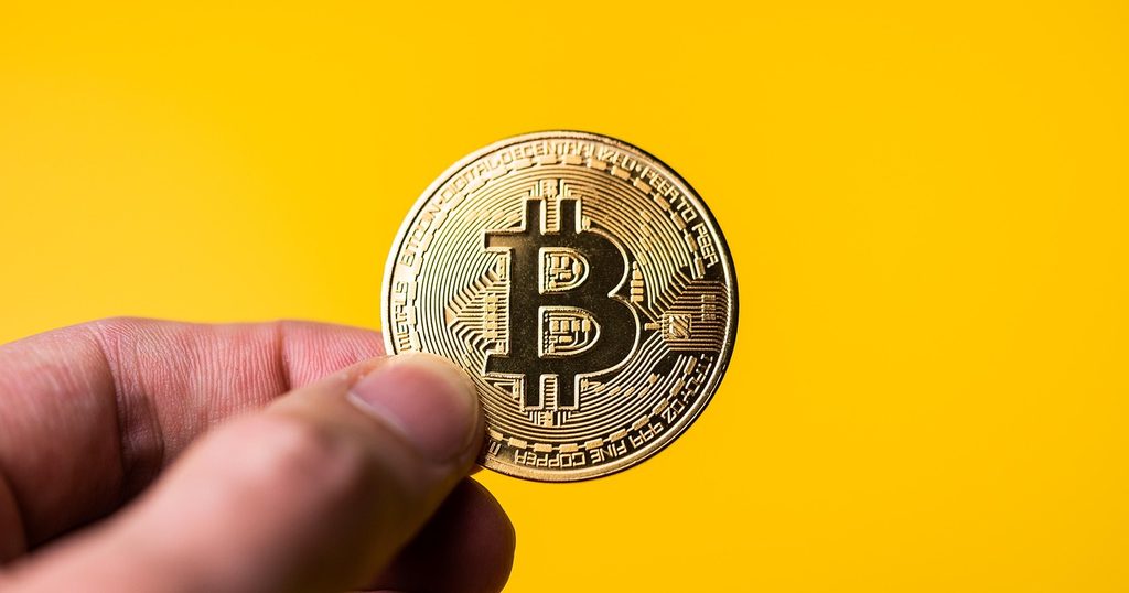 黄色い背景の前で指先に持った、金色のビットコインの記念コイン